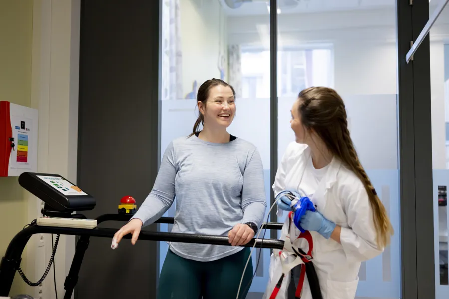 En dame i hvit labfrakk som smiler til en dame som trener på tredemølle