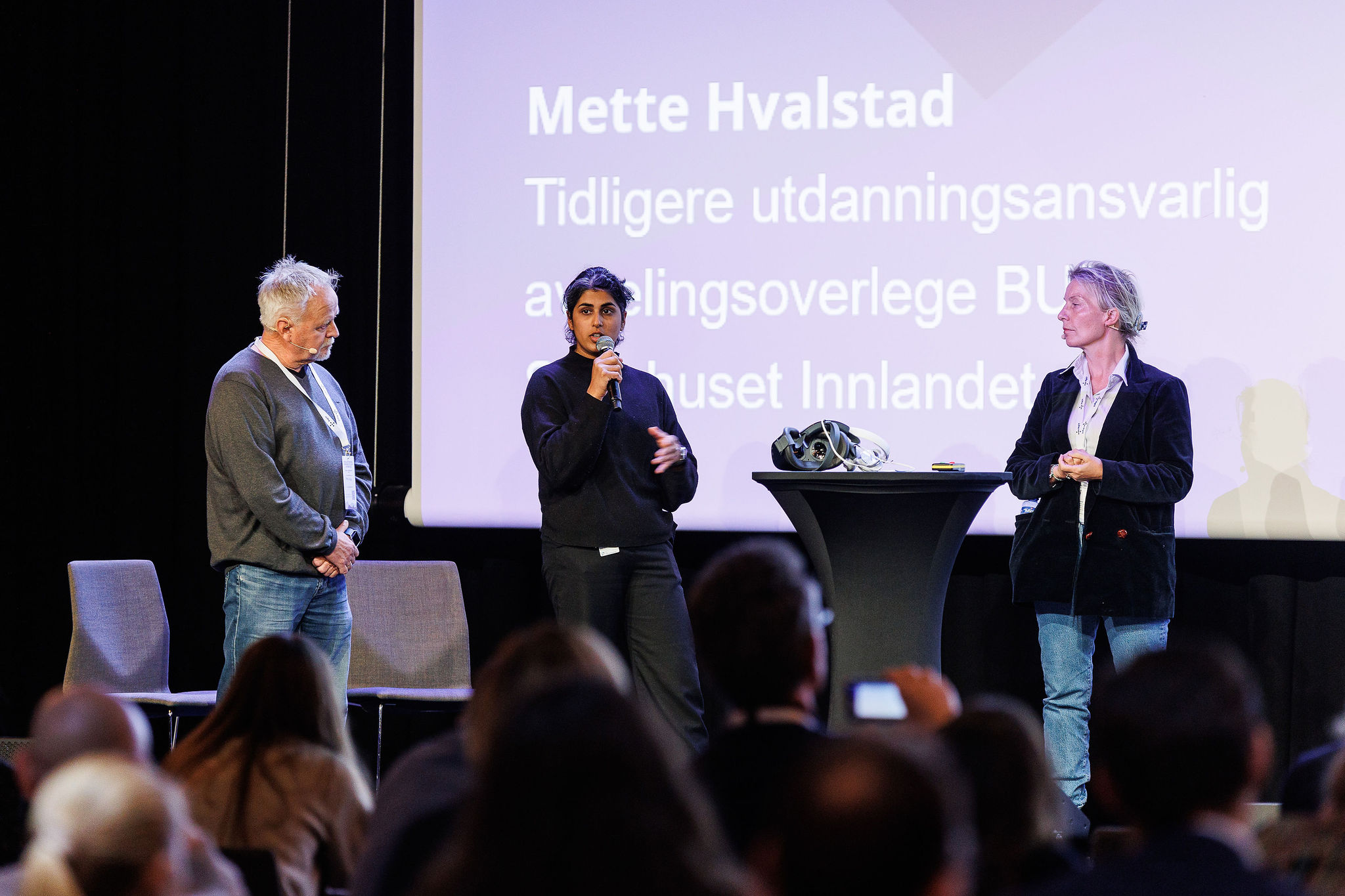 Tre personer står på scenen og snakker sammen. Den i midten holder en mikrofon og har ordet