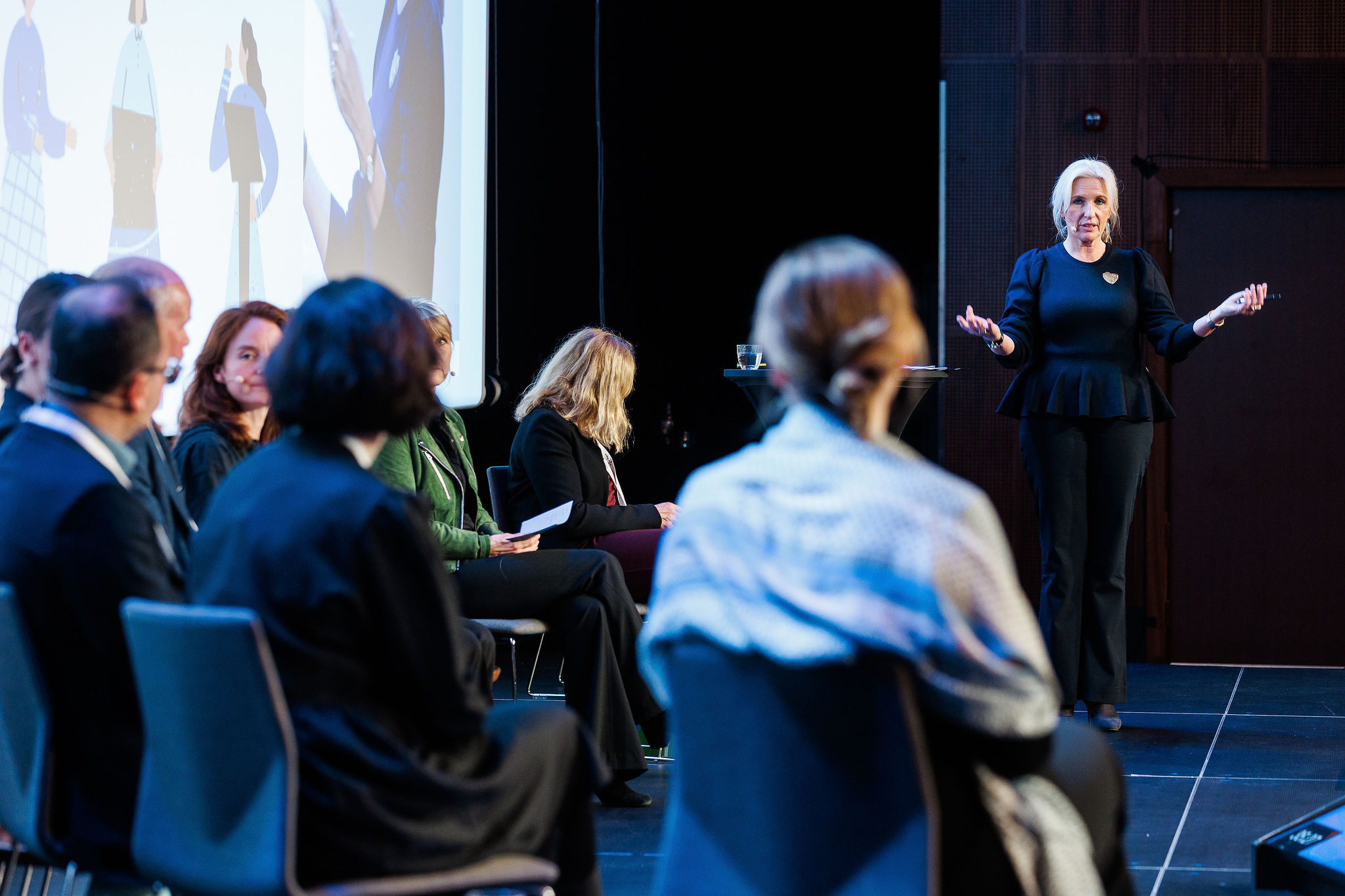 Personer som sitter på stoler på scenen, noen ser vi bakfra andre fra siden. En person står til høyre og snakker