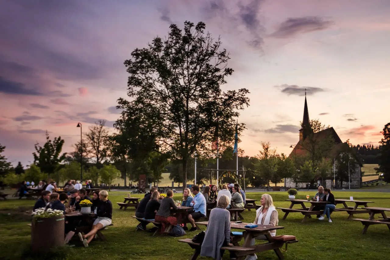 Mennesker som sitter på benker med bord på Stiklestad, med vakker himmel i bakgrunnen